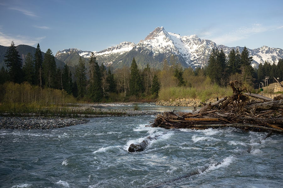 Whitehorse Mountain North Cascades Darrington WA Sauk River Snow-capped mountain rising above a forested valley with a fast-flowing river in the foreground, scattered driftwood along the bank, and a bridge visible to the right under a clear blue sky.