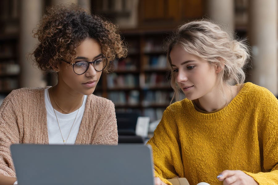Two young women sit side by side at a table, focused on a laptop, collaborating in a library or study space with bookshelves in the background.