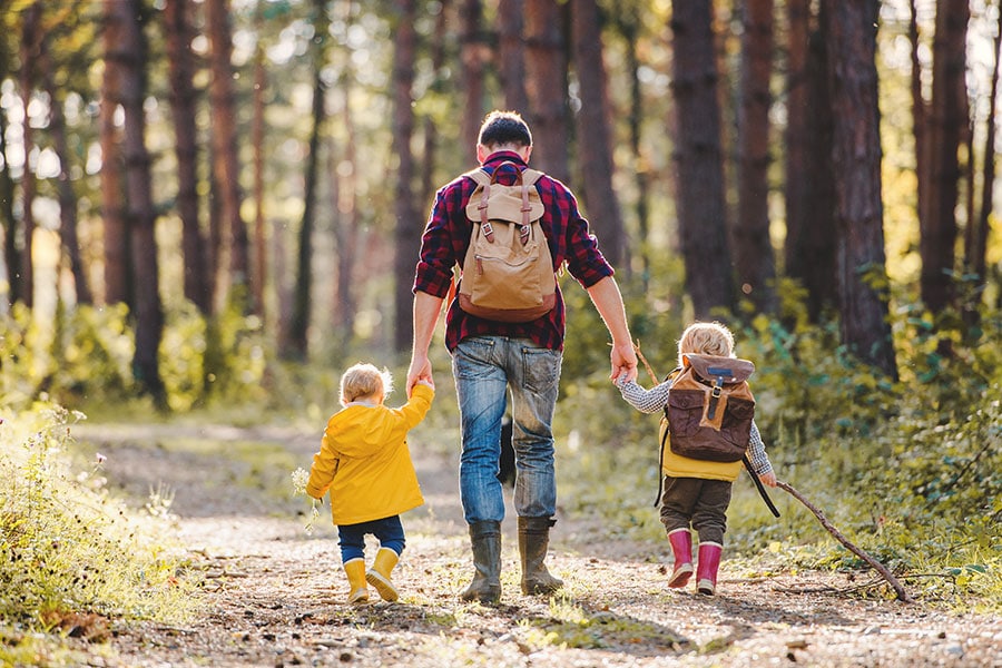 An adult walks along a forest trail holding hands with two young children, all wearing backpacks, enjoying a sunny outdoor hike.