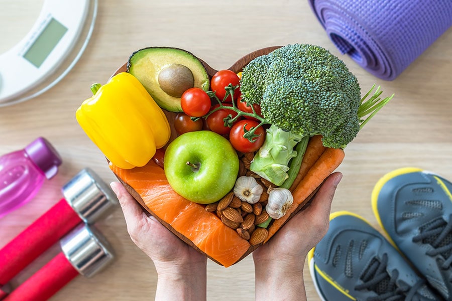 Hands holding a heart-shaped bowl filled with healthy foods like salmon, avocado, broccoli, apple, carrots, tomatoes, nuts, and garlic, surrounded by fitness items including dumbbells, a water bottle, a yoga mat, and sneakers.