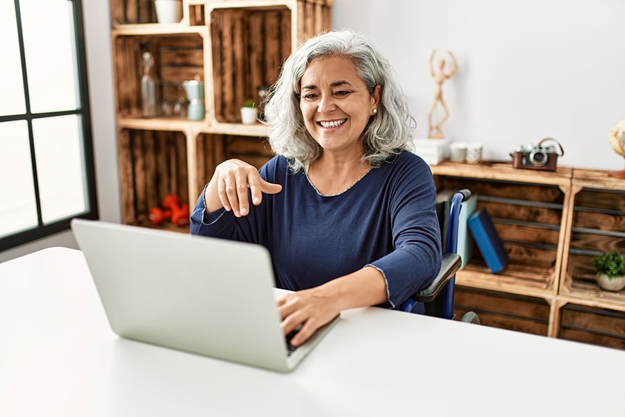 A smiling older woman with gray hair sits at a desk, using a laptop and gesturing with her hand, as if engaged in a video call, in a cozy home office with wooden shelves in the background.