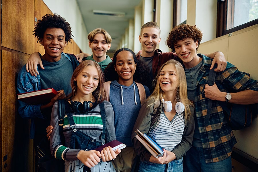 A group of smiling teenagers stand together in a school hallway, holding books and wearing backpacks, posing for a photo.