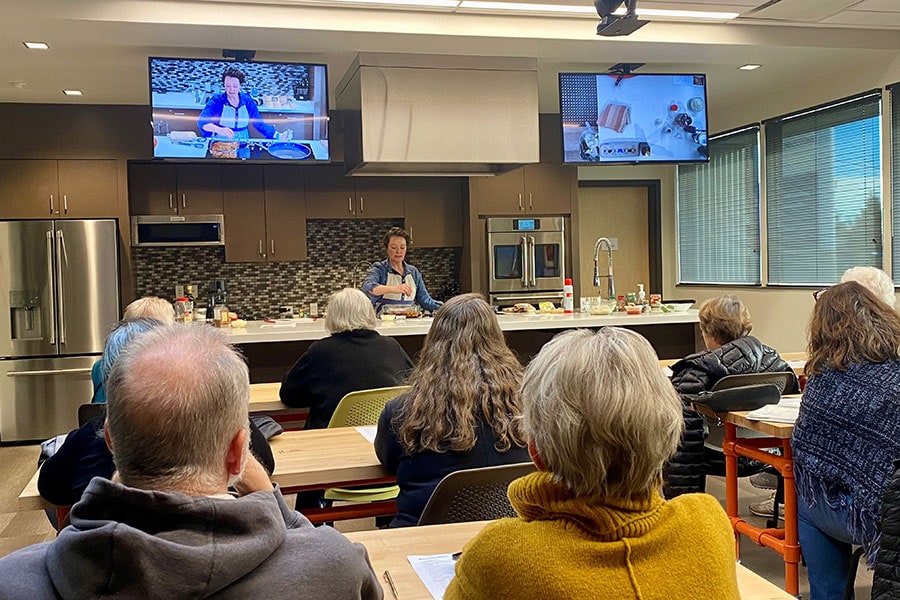 A group of adults sit at tables watching a cooking demonstration in a modern kitchen classroom, where an instructor prepares food at the front while the process is shown on overhead screens.