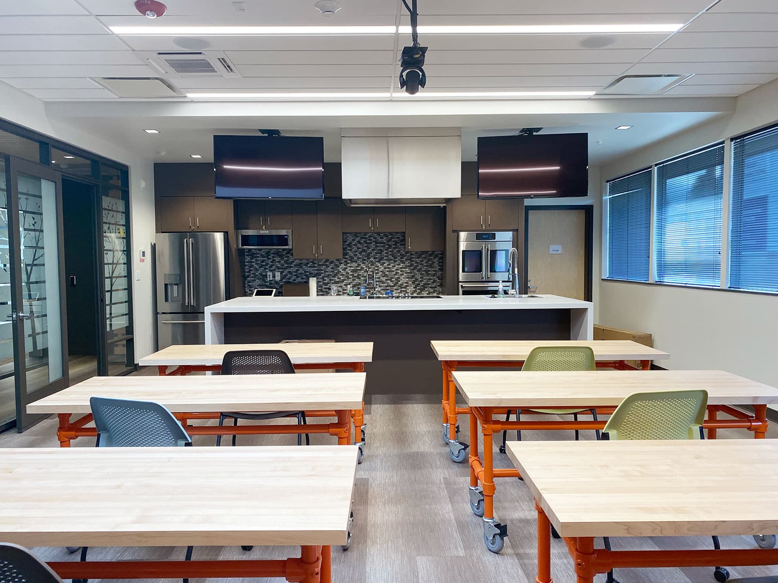 An empty kitchen classroom with rows of tables and chairs facing a demonstration counter, stainless steel appliances, and two overhead screens at the front.
